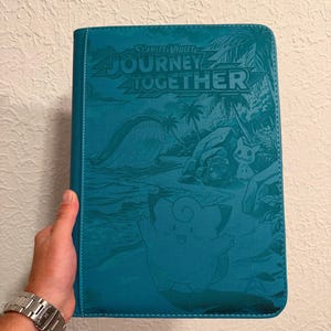 Könnte beinhalten: Ein rechteckiger Sammelkartenordner in Blaugrün mit einem geprägten Design aus verschiedenen Kreaturen und Palmen. Der Text "SCARLET & VIOLET JOURNEY TOGETHER" ist oben aufgedruckt. Der Einband hat eine strukturierte Oberfläche und genähte Kanten.