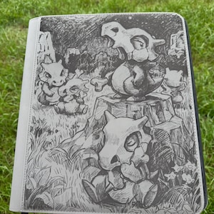 Könnte beinhalten: Ein grauer Reißverschlussordner mit einer detaillierten schwarz-weißen Skizze mehrerer Cubone-Charaktere in einer Graslandschaft. Ein Cubone sitzt auf einem Baumstumpf, während andere im umliegenden Gras und Gebüsch positioniert sind.