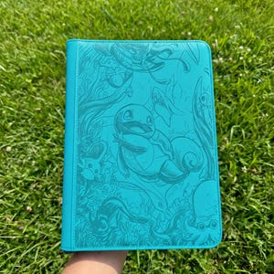 Könnte beinhalten: Ein rechteckiger türkisfarbener Ordner mit einem geprägten Grafikdesign verschiedener Wasserlebewesen, darunter eine zentrale Figur, die einer Schildkröte ähnelt, umgeben von Algen, Fischen und anderen Meereslebewesen auf dem Vorderdeckel.