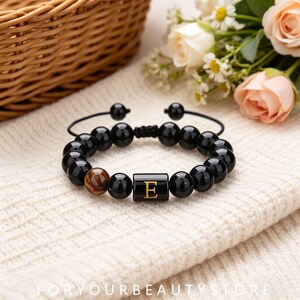 Könnte beinhalten: Ein Perlenarmband mit glatten schwarzen Kugeln, einer braunen Tigeraugenperle und einer zentralen zylindrischen schwarzen Perle mit einem goldenen Buchstaben E. Das Armband hat einen verstellbaren Kordelverschluss mit zwei kleinen schwarzen Perlen.
