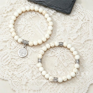 Könnte beinhalten: Zwei elastische Armbänder aus runden, cremeweißen Perlen mit brauner Maserung. Ein Armband hat einen silberfarbenen runden Anhänger mit Baummotiv. Das andere Armband enthält drei silberfarbene zylindrische Zwischenstücke mit Mustern.
