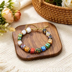 Könnte beinhalten: Ein Armband mit bunten, quadratischen Perlen in verschiedenen Mustern, darunter florale und gestreifte Designs, unterbrochen von kleinen goldfarbenen Metallzwischenstücken. Das Armband verfügt über eine Verlängerungskette und einen Karabinerverschluss.
