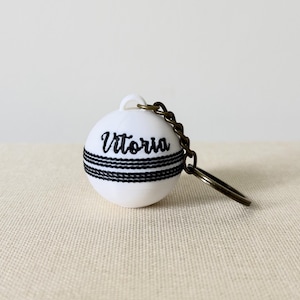 May include: A white spherical keyring featuring a black stitched seam pattern resembling a cricket ball. The name Vitoria is printed in black script on the upper half of the sphere. A dark bronze-toned metal chain and ring are attached to the top.