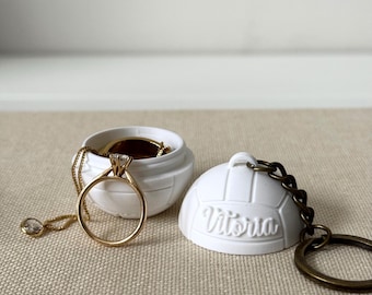 May include: A white, spherical, volleyball-shaped container with a detachable lid featuring the name Vitoria in raised script. The open container holds a gold-toned ring and a delicate gold chain, while the lid is attached to a bronze keyring.