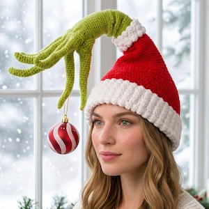 May include: A red and white crocheted Santa hat featuring an attached long, green, textured, hand-shaped extension that reaches out to hold a small, round, red and white striped Christmas ornament hanging from the fingers.