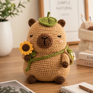 Peut inclure: Une peluche capybara en crochet beige avec un museau marron foncé, des yeux fermés et des joues roses. Elle porte une feuille verte sur la tête, un petit sac vert en bandoulière et tient un tournesol jaune au centre marron dans sa patte.