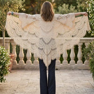 Può includere: Uno scialle in pizzo color crema caratterizzato da un intricato motivo a ventaglio smerlato con delicati dettagli traforati. Il capo è indossato sulle spalle, estendendosi verso l'esterno con maniche ampie e fluide e un orlo a punta.