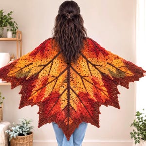 May include: A crochet shawl shaped like a large maple leaf, featuring a gradient of autumn colors including deep burgundy, vibrant orange, golden yellow, and hints of brown, with a detailed central vein and jagged, pointed edges.