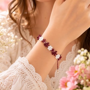 May include: A delicate bracelet featuring a repeating pattern of small, six-petaled flowers in alternating deep burgundy and crisp white thread. The floral motifs are connected in a continuous chain, finished with a thin dangling cord at the end.