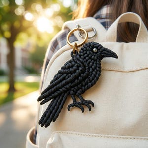 May include: A black macrame raven keychain featuring a detailed knotted cord design, a small silver-toned bead for the eye, and a circular brass-colored metal ring for attachment, secured to the top of a cream-colored canvas backpack.