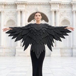 May include: A dark charcoal gray crochet capelet featuring an intricate layered feather design that extends outward from the shoulders and chest. The garment is constructed with detailed stitch patterns resembling bird wings, creating a textured, pointed silhouette that drapes over the upper body.