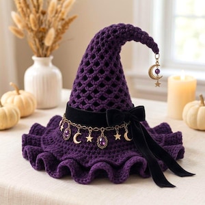 Puede incluir: Un sombrero de ganchillo de color morado intenso con un ala ancha y ondulada y una parte superior puntiaguda. El sombrero presenta una cinta de terciopelo negro atada en lazo, adornada con una cadena dorada con estrellas, lunas y gemas.