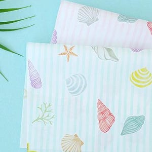 Puede incluir: Dos piezas de tela blanca dobladas con un patrón repetitivo de conchas marinas, estrellas de mar y corales coloridos. La tela tiene finas rayas verticales de color azul claro y una textura lisa, mostrando varias formas de conchas en rosa, amarillo y azul.