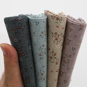 May include: A hand holds four rolled fabric pieces. The fabrics are patterned with small floral designs in shades of gray, blue, beige, and lavender. The floral patterns include small red and brown flowers.