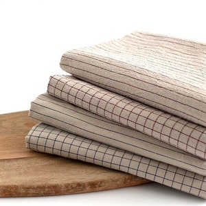 May include: Four beige and brown checkered fabric napkins stacked on a wooden cutting board. The napkins have different sized squares and are made of a soft, textured material.