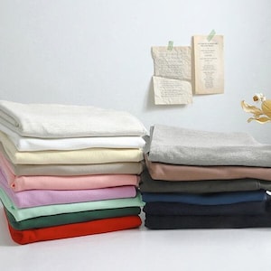 Pode incluir: Uma pilha de tecido dobrado em várias cores, incluindo bege, branco, creme, rosa, lavanda, verde e vermelho. O tecido está disposto em duas pilhas sobre uma superfície branca.