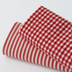May include: Two folded fabric pieces. One is red and white gingham, the other is red and white striped. The fabrics are layered on a white background. The fabrics are likely for home decor or crafting.