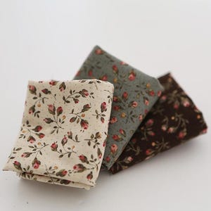 May include: Three folded fabric squares with floral patterns. The fabrics feature a cream, gray-green, and brown background, each adorned with delicate pink and red rose-like flowers and green leaves. The fabrics are stacked on a white surface.