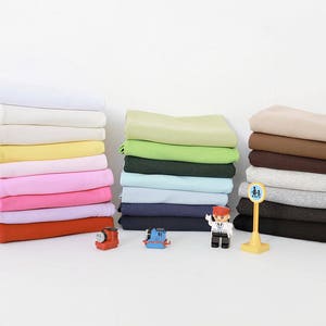 Puede incluir: Una pila de muestras de tela en varios colores, incluyendo blanco, amarillo, rosa, rojo, verde, marrón y gris. Las muestras de tela están dispuestas en una fila sobre una superficie blanca. También hay dos trenes de juguete y una persona de juguete en la superficie.