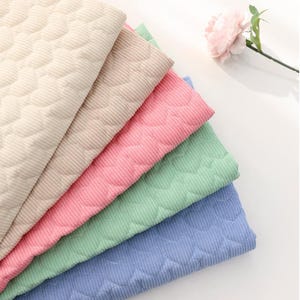 Op de afbeelding: Een stapel kleurrijke corduroy stofstalen in beige, roze, groen en blauw. Elk stuk heeft een gewatteerd patroon. Een roze bloem met groene stengel staat op de achtergrond.