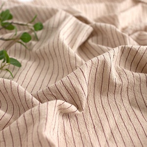 May include: Close-up of a beige fabric with thin, red stripes. The fabric is wrinkled and has a soft, textured appearance.