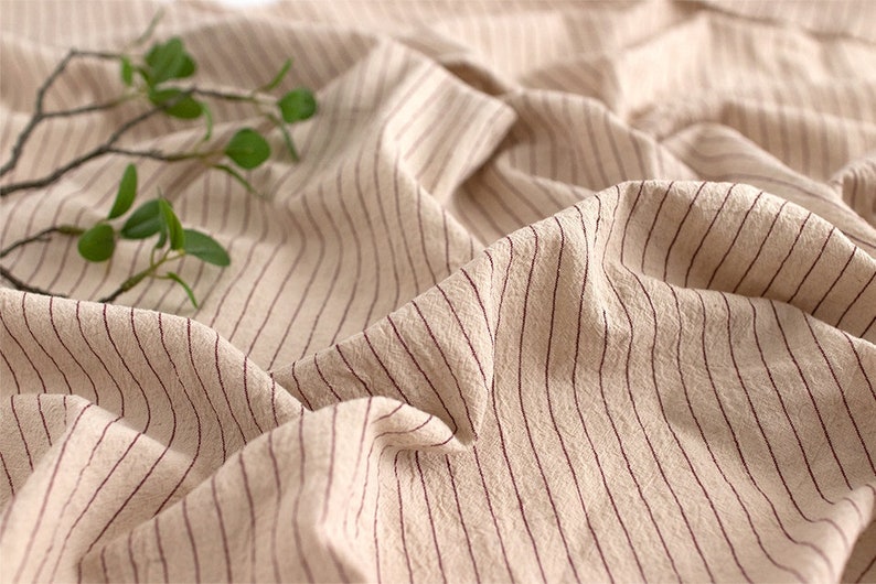 May include: Close-up of a beige fabric with thin, red stripes. The fabric is wrinkled and has a soft, textured appearance.