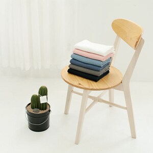 Op de afbeelding: Een stapel van zes vierkante stukken katoenen stof in verschillende kleuren, waaronder wit, roze, blauw, grijs en zwart, liggen op de zitting van een houten stoel. De stoel is wit met een houten zitting en rugleuning. Op de voorgrond staat een cactusplant in een pot.