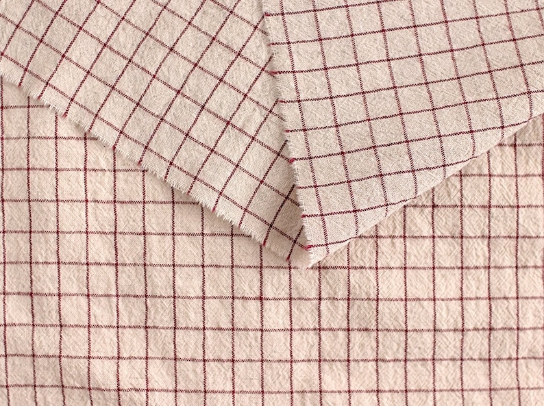 May include: Two pieces of beige fabric with a red checkered pattern. The fabric is folded over itself, showing the pattern on both sides.