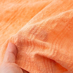 May include: Close-up of a soft, peach-colored fabric with a subtle, textured pattern. The fabric is slightly wrinkled and has a light, airy feel.