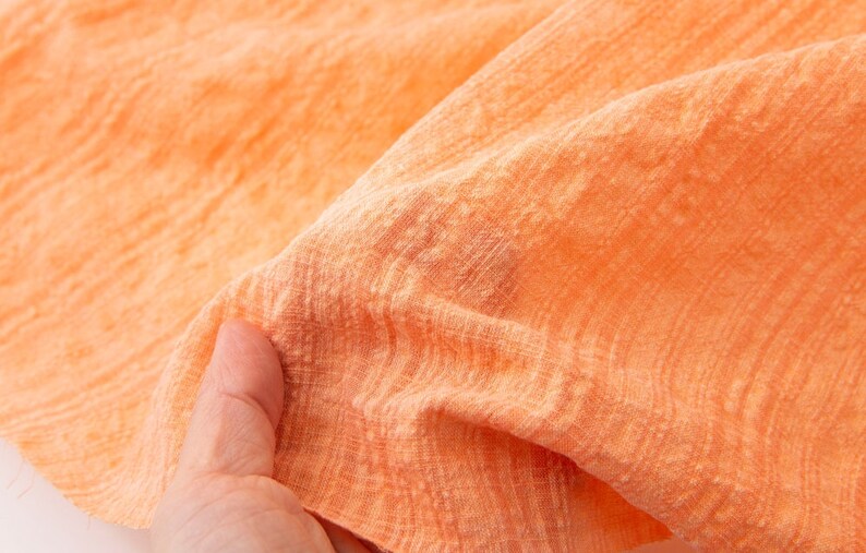 May include: Close-up of a soft, peach-colored fabric with a subtle, textured pattern. The fabric is slightly wrinkled and has a light, airy feel.
