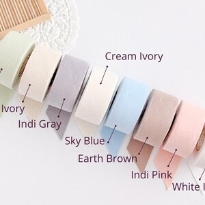 May include: A collection of eight rolls of decorative ribbon in various pastel colors, including green tea, natural ivory, indi gray, sky blue, earth brown, indi pink, cream ivory, and white ivory. Each roll is labeled with its color name.