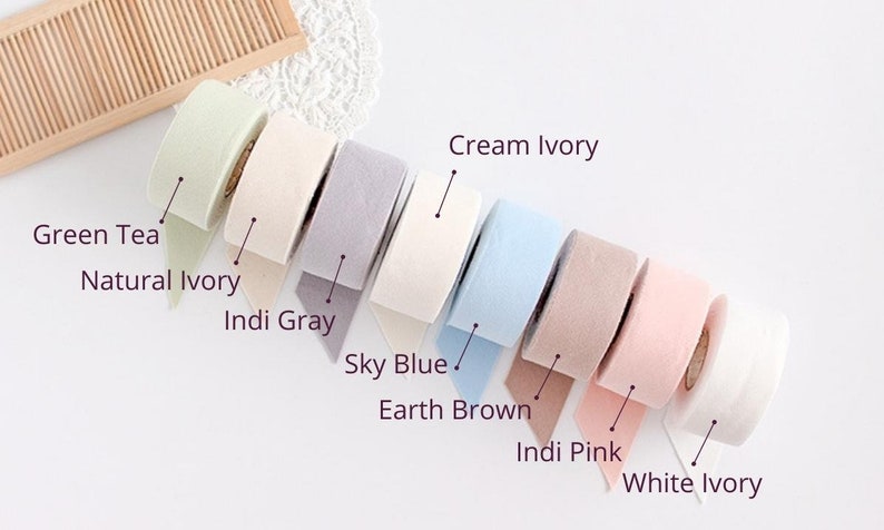 May include: A collection of eight rolls of decorative ribbon in various pastel colors, including green tea, natural ivory, indi gray, sky blue, earth brown, indi pink, cream ivory, and white ivory. Each roll is labeled with its color name.