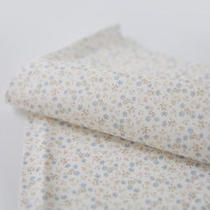 Puede incluir: Una tela doblada con un delicado estampado floral. La tela es principalmente blanquecina, con pequeños diseños florales y de hojas azules, rosas y marrones dispersos por la superficie. La tela parece suave y ligera.