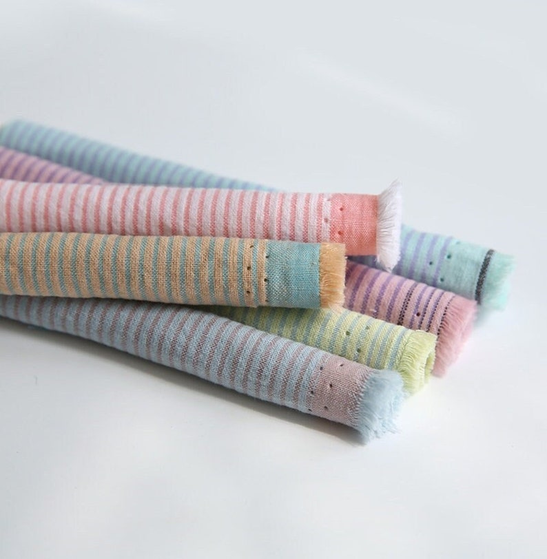 May include: A stack of five striped fabric swatches in pastel colors, including pink, blue, green, and yellow. The fabric has a textured, woven look and a frayed edge.