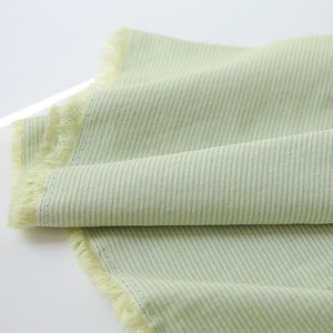 May include: A light green fabric with thin white stripes. The fabric has a frayed edge.