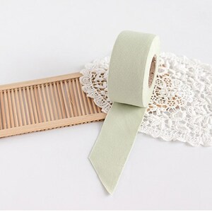 May include: A roll of light green washi tape with a portion unrolled, revealing its adhesive side. The tape sits on a white lace doily and a small, light brown bamboo tray. The background is white.