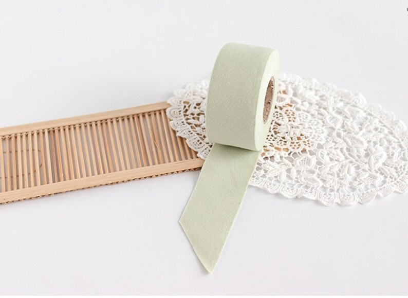 May include: A roll of light green washi tape with a portion unrolled, revealing its adhesive side. The tape sits on a white lace doily and a small, light brown bamboo tray. The background is white.