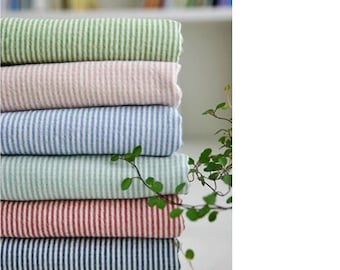 May include: A stack of five striped fabric swatches in various colors, including green, pink, blue, light green, and red. The fabric is folded neatly and arranged on a white surface. A green plant is in the background.