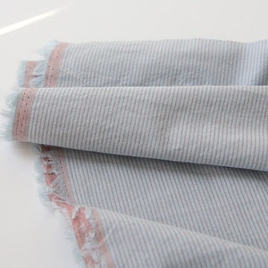 May include: A close-up of a light blue and pink striped fabric with a frayed edge. The fabric has a subtle texture and a soft, casual look.