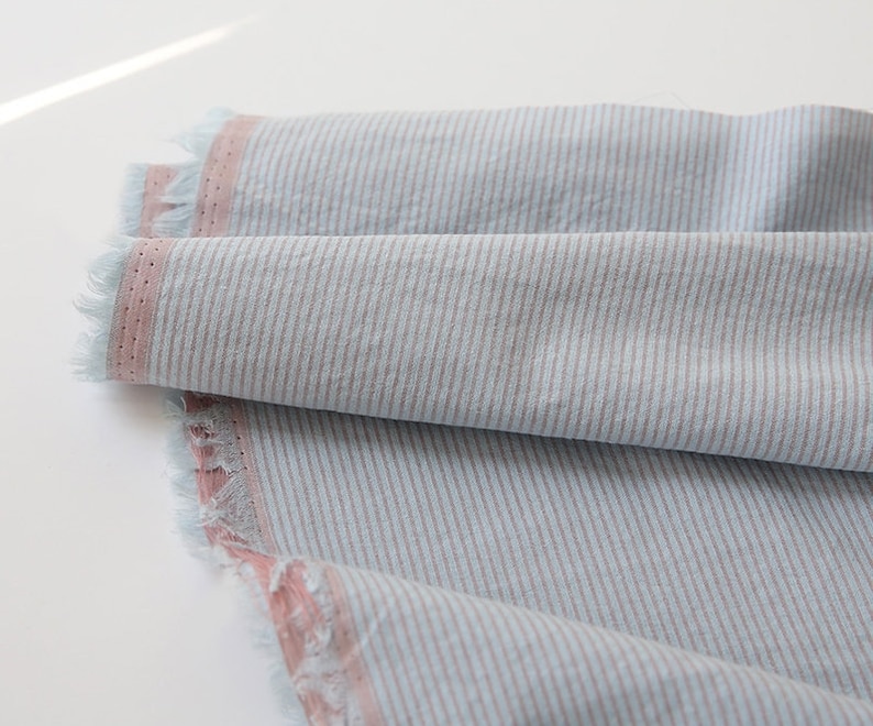 May include: A close-up of a light blue and pink striped fabric with a frayed edge. The fabric has a subtle texture and a soft, casual look.