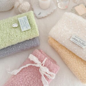 May include: A collection of soft, textured items in pastel colors. Includes folded fabric pieces in light green, gray, and pink, some with tags that read "Made in Dreamland" and "Atelier De Tout". A small white candle and decorative elements are also present.
