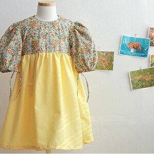 May include: A yellow and floral print dress with puffed sleeves and a tie at the wrist. The dress has a yellow skirt and a floral print bodice.