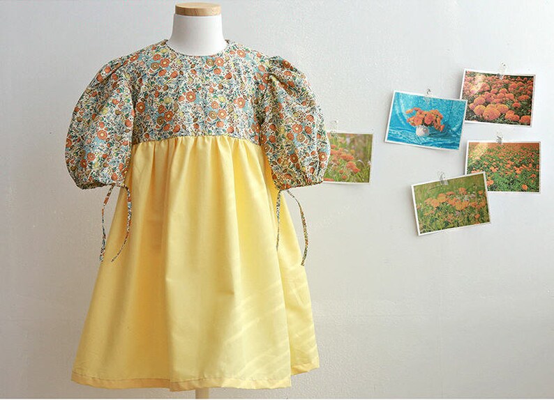May include: A yellow and floral print dress with puffed sleeves and a tie at the wrist. The dress has a yellow skirt and a floral print bodice.