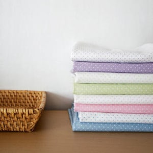 May include: A stack of folded fabric in various pastel colors, including white, lavender, green, pink, and blue. Each piece of fabric has a repeating pattern of small white dots. A woven basket sits to the left on a wooden surface.
