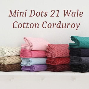 Pode incluir: Uma pilha de oito amostras de tecido de veludo cotelê de algodão de diferentes cores com pequenos pontos brancos. O tecido é dobrado ao meio e empilhado em uma prateleira branca. O texto "Mimi Dots 21 Wale Cotton Corduroy" está escrito em marrom na parte superior da imagem.