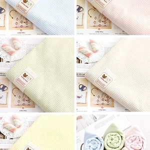 May include: Four different striped fabric swatches with a teddy bear logo. The fabric swatches are in blue, pink, green, and yellow. The swatches are rolled up in a basket.