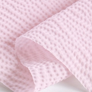 May include: Close-up of a soft, light pink fabric with a textured, waffle-like pattern. The fabric is folded, revealing the interior and creating shadows that highlight the texture. The material appears lightweight and airy.