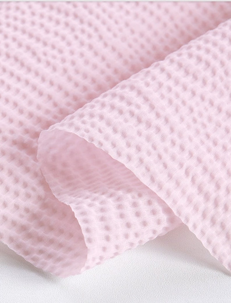 May include: Close-up of a soft, light pink fabric with a textured, waffle-like pattern. The fabric is folded, revealing the interior and creating shadows that highlight the texture. The material appears lightweight and airy.