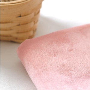 May include: A close-up of a soft, pink fabric with a velvety texture. The fabric is folded, revealing its thickness and subtle variations in color. A small, woven basket is partially visible in the background, adding a touch of rustic charm.