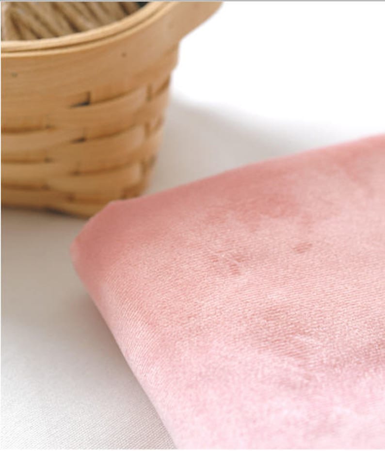 May include: A close-up of a soft, pink fabric with a velvety texture. The fabric is folded, revealing its thickness and subtle variations in color. A small, woven basket is partially visible in the background, adding a touch of rustic charm.